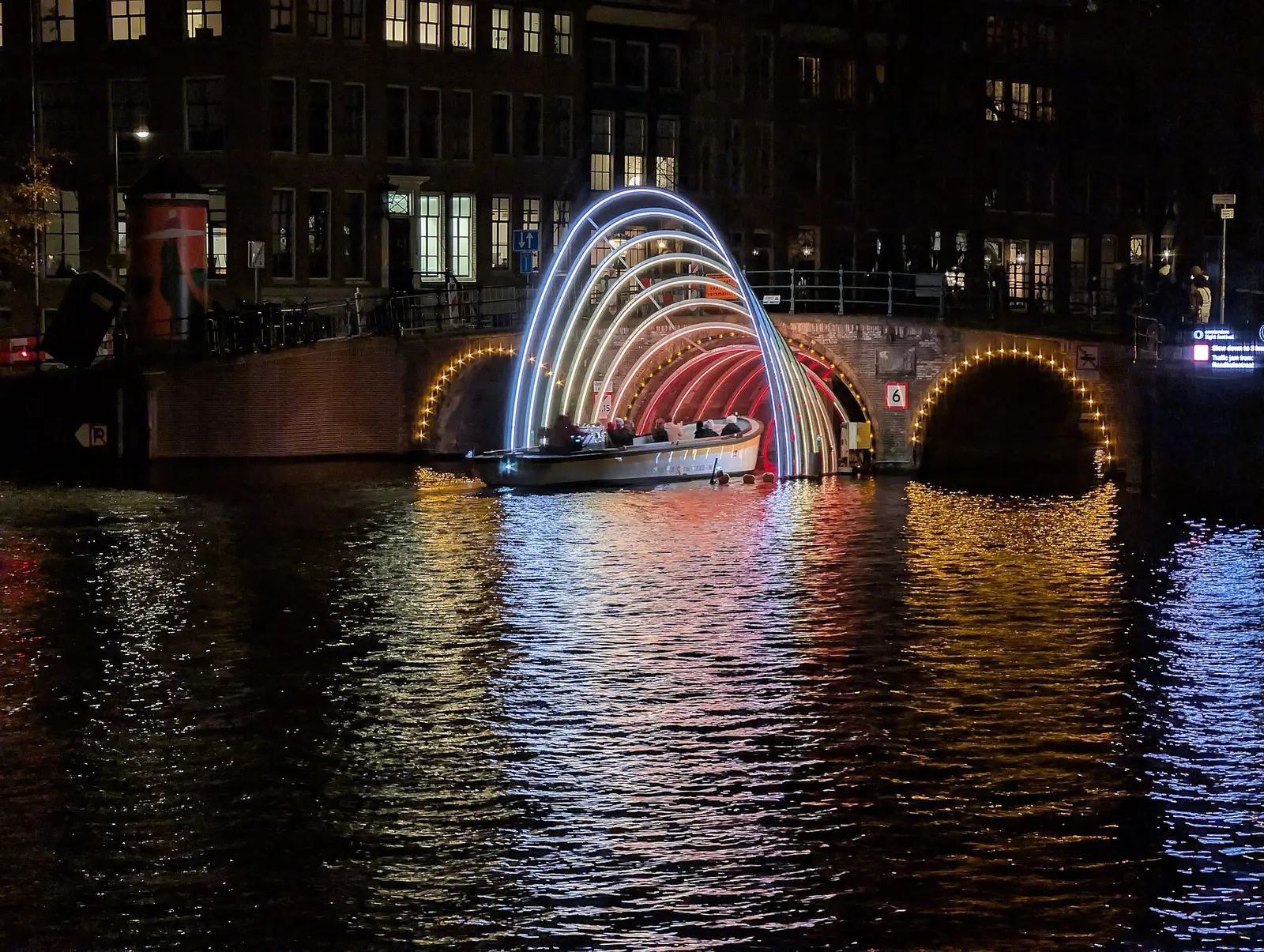A canal boat approaches the lit rainbow arch artwork Solstice under a bridge at night during Amsterdam Light Festival 2024
