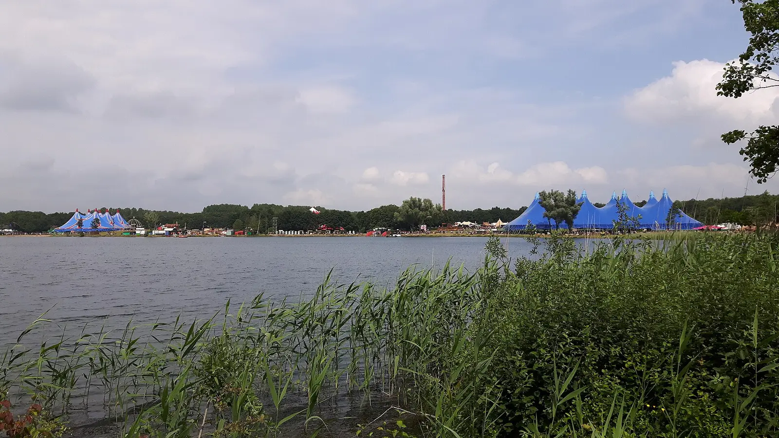 Blue festival tents on the far shore of a lake with reeds in the foreground at Down The Rabbit Hole in 2016