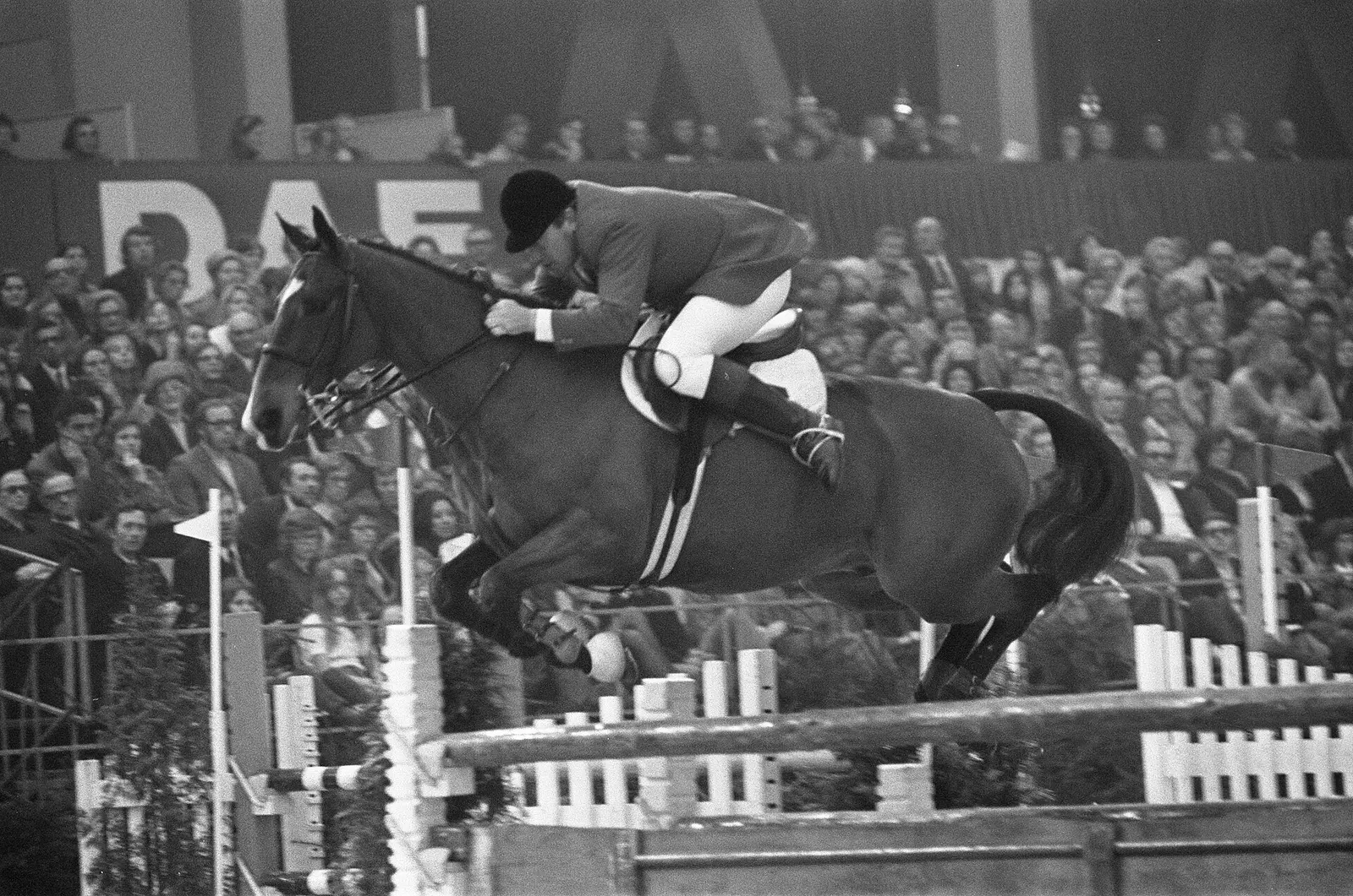 A rider clears a fence on a horse in front of a full crowd at the Jumping Amsterdam show in the RAI in black and white