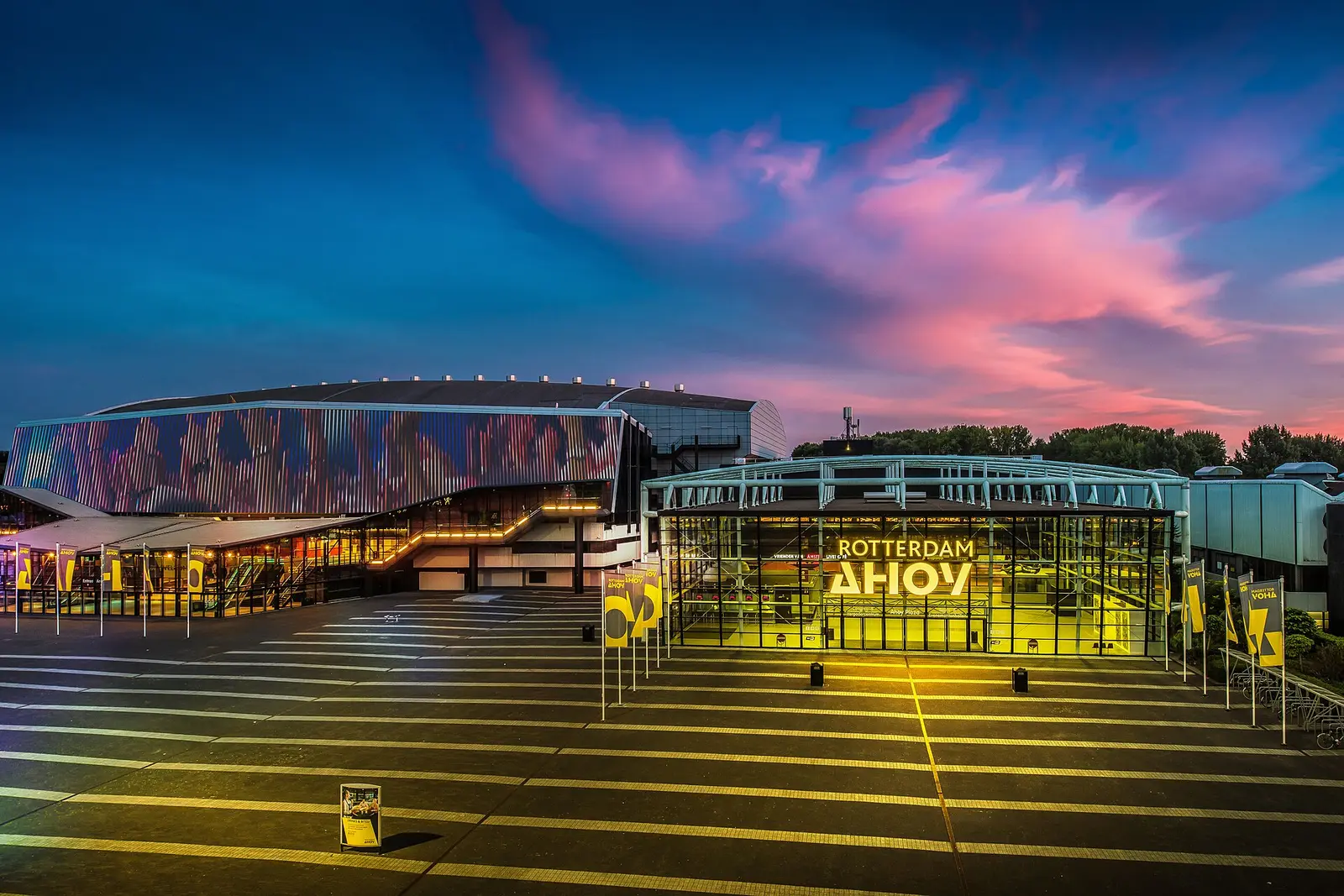 The entrance and arena of Rotterdam Ahoy at dusk with a lit sign and pink clouds